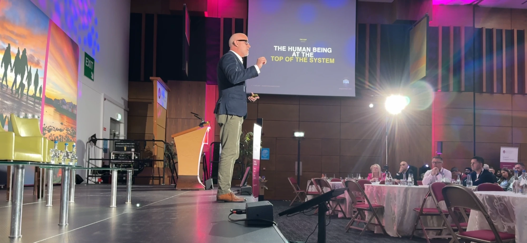 Angus Nelson on stage at a conference, presenting a slide titled The Human Being at the Top of the System, with attendees seated at tables